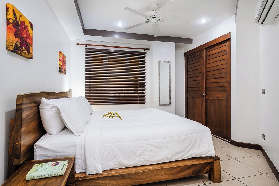 Une chambre à coucher avec un lit en bois, du linge de lit blanc, un ventilateur de plafond, une fenêtre avec des stores et une armoire en bois.