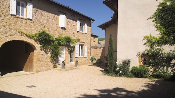 A stone building with a large arched entrance, a second-story window with shutters, and a small garden area with potted plants and a tall tree. - Beaujolais house with charm, pool, fireplace (Gleizé)