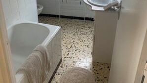Ein Badezimmer mit einer weißen Badewanne, einem Waschbecken und einer Duschkabine. Ein Fenster sorgt für natürliches Licht, und ein Handtuch hängt an der Wand.