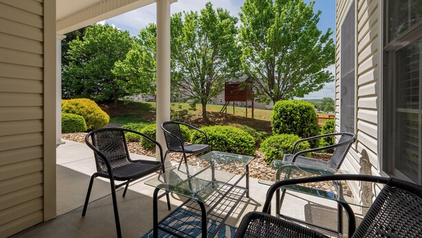 A covered patio with outdoor furniture, a glass table, and a view of a well-maintained garden and trees.