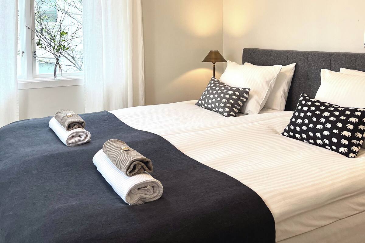 A neatly made bed with a black and white patterned pillow, a rolled towel, and a lamp on a bedside table.