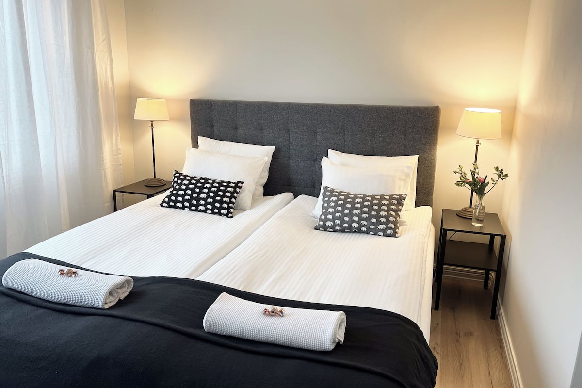 A double bed with white linens, two pillows with a black and white pattern, and two rolled towels. There are two bedside tables with lamps and a vase with flowers.