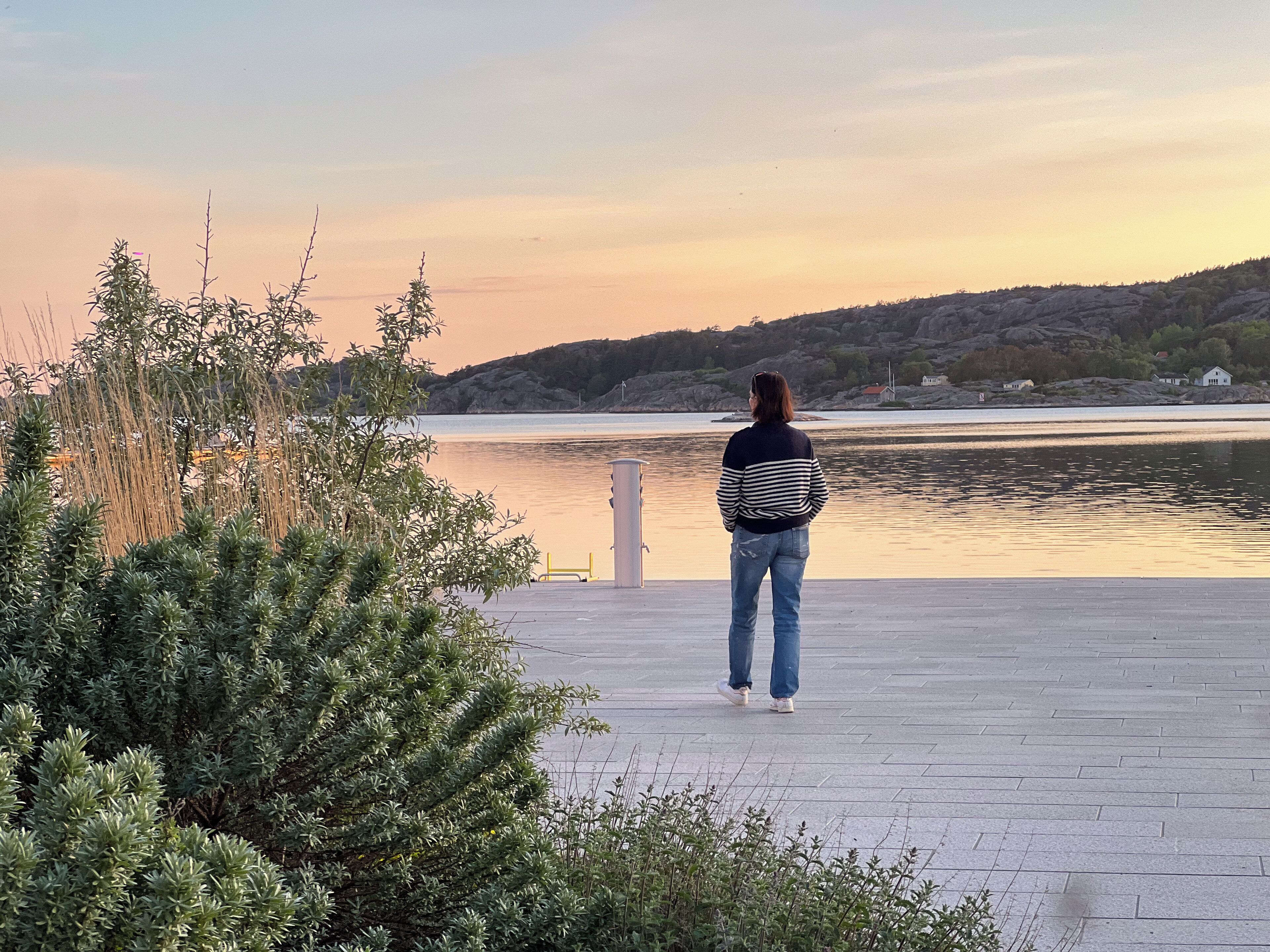 A person standing on a paved area overlooking a body of water with hills in the background.