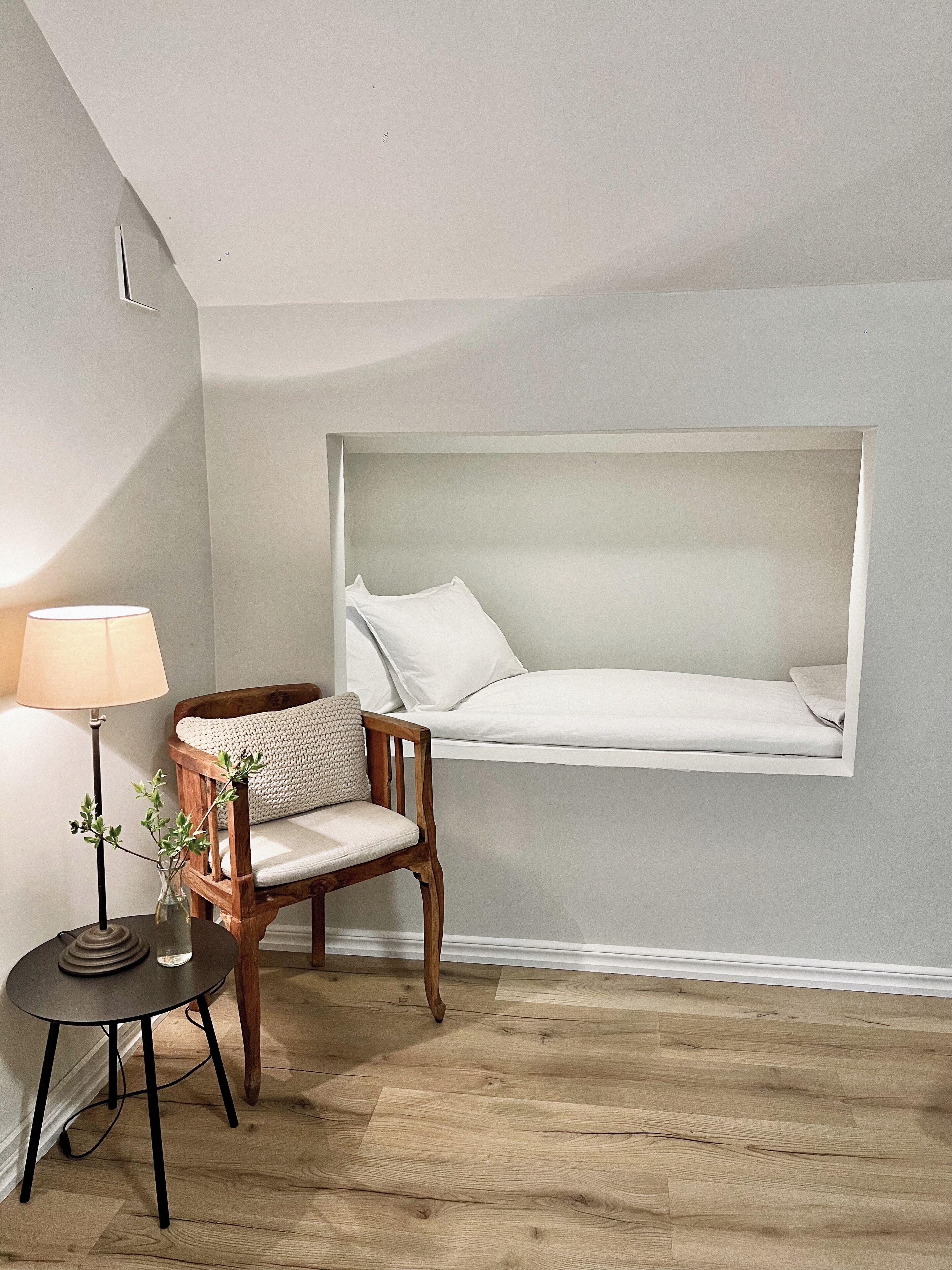 Un dormitorio con cama empotrada junto a la ventana, una silla de madera, una mesita con lámpara y un florero con plantas.