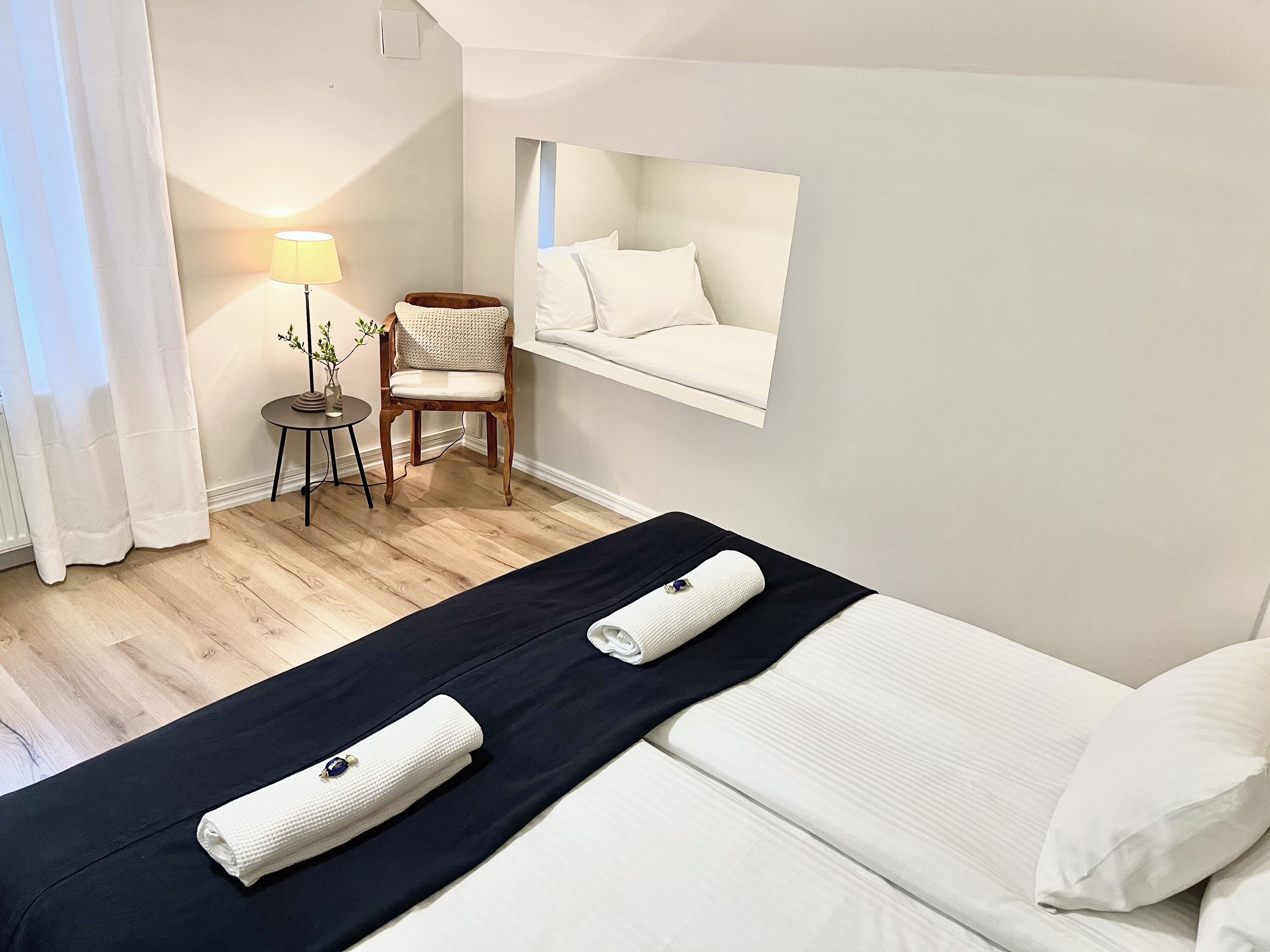 A hotel room with a bed, a chair, a small table with a lamp, and a window with a view of the sky.