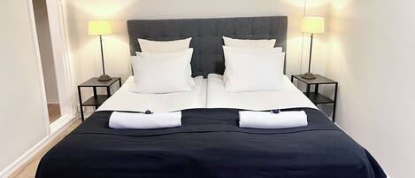 Una cama doble con cabecera oscura, dos mesitas de noche con lámparas y una colcha negra.