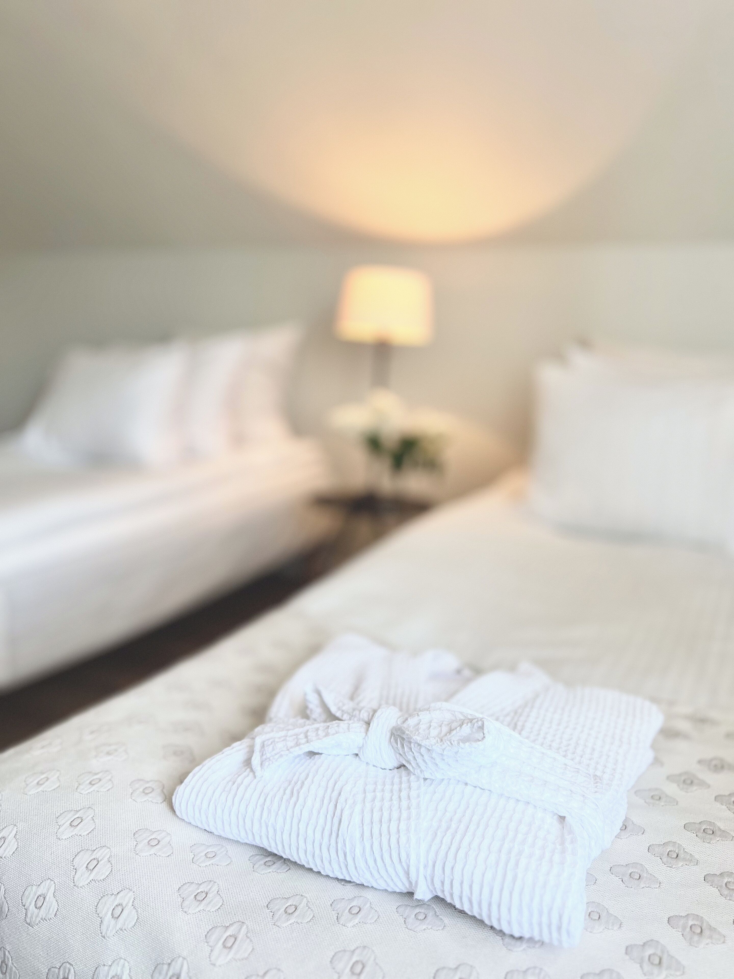 A hotel room with two beds, a nightstand with a lamp, and a neatly folded white robe on the bed.