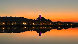 Un lac paisible au coucher du soleil, avec une petite colline sur laquelle se dresse un bâtiment imposant illuminé de lumière violette.