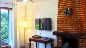 Una sala de estar con chimenea de ladrillo, televisor montado en la pared, una mesa con sillas y una ventana con cortinas.