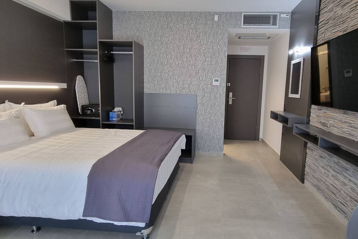 A modern hotel room with a large bed, built-in wardrobe, a flat-screen TV mounted on the wall, and a small seating area.