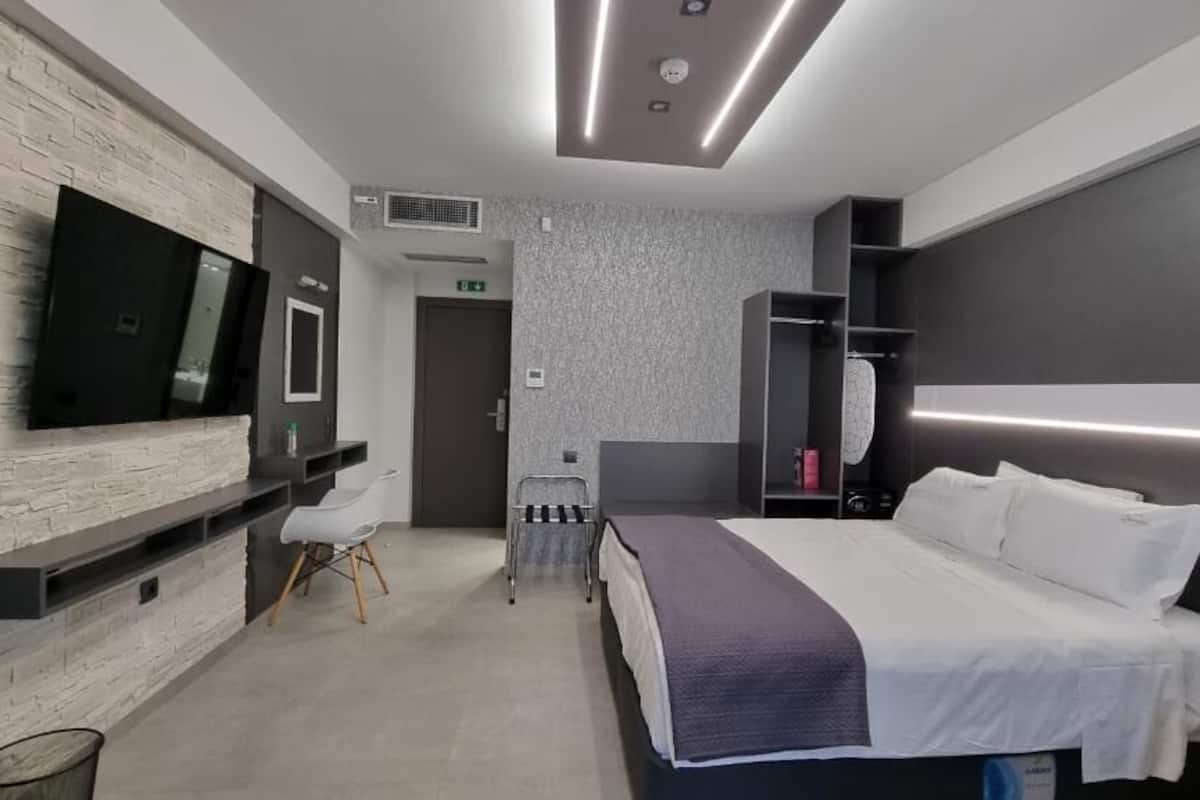 A modern hotel room with a large bed, a flat-screen TV mounted on a textured wall, a seating area with a chair, and a small table.
