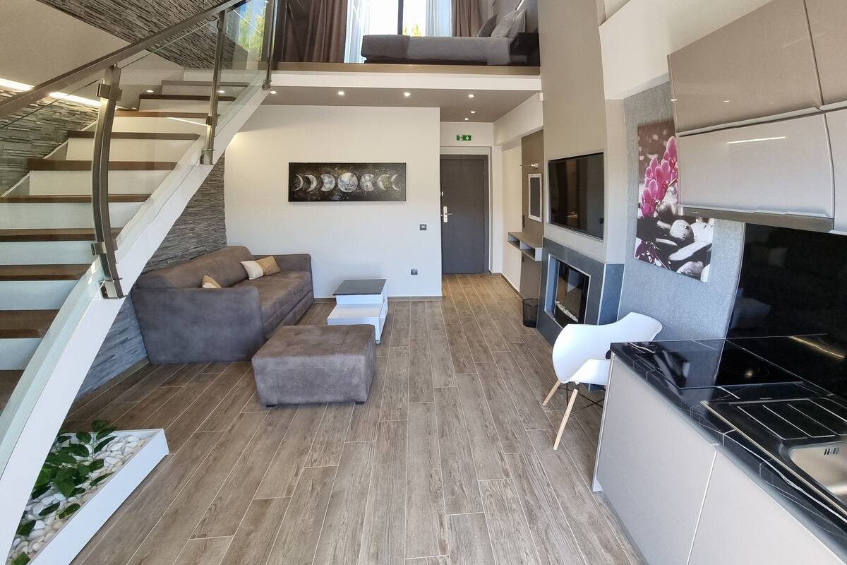 A spacious living area with a staircase, a sofa, ottoman, and a kitchen area with a TV and fireplace.