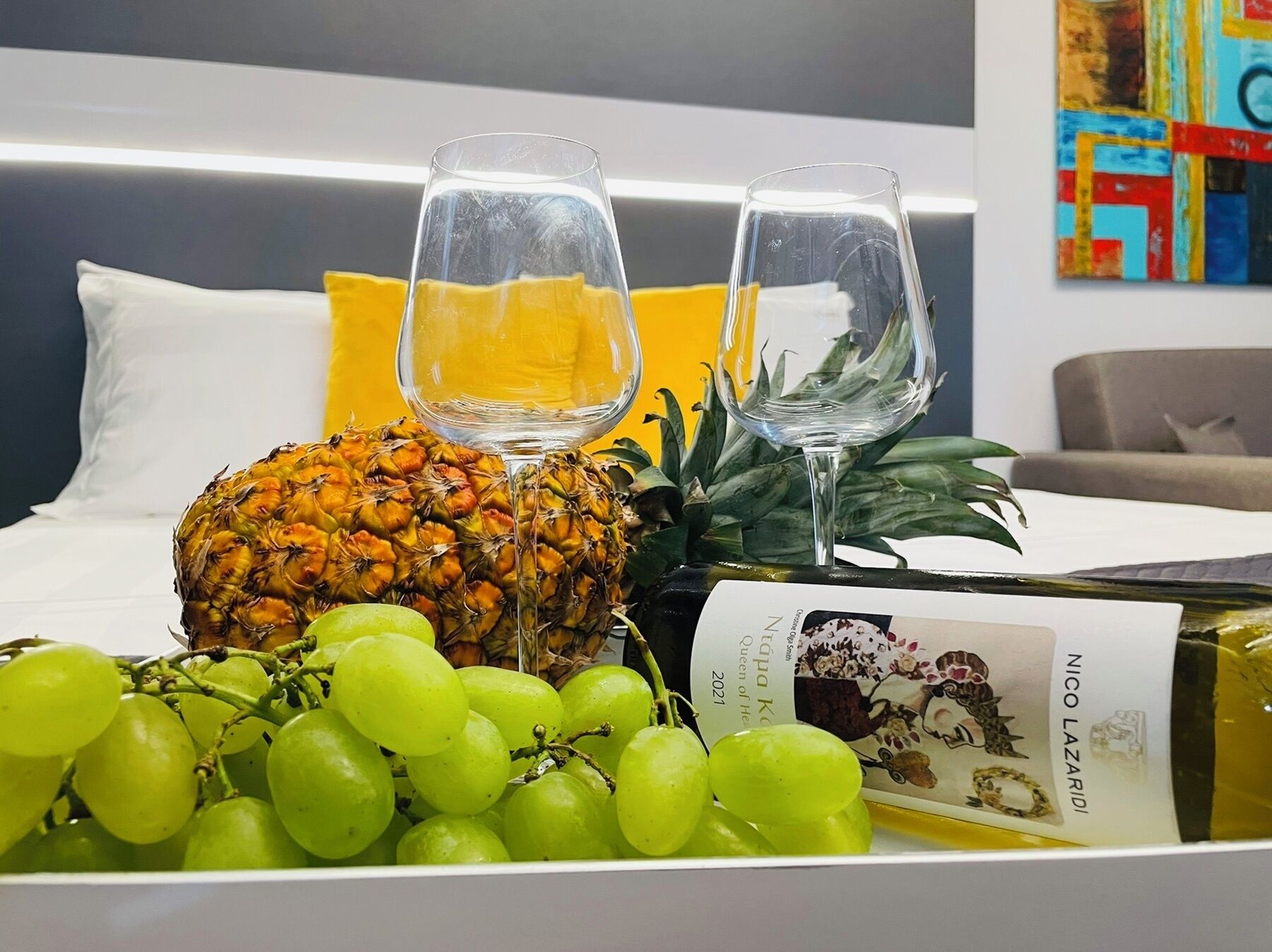 A bed with white pillows and a yellow accent pillow, a pineapple, a bunch of green grapes, two wine glasses, and a bottle of wine on a tray.