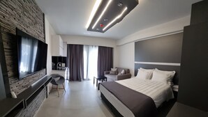 A modern hotel room with a large bed, a flat-screen TV mounted on a textured wall, a desk with a chair, a sofa, and a large window with curtains. - LUXSUITES (Pangaio)