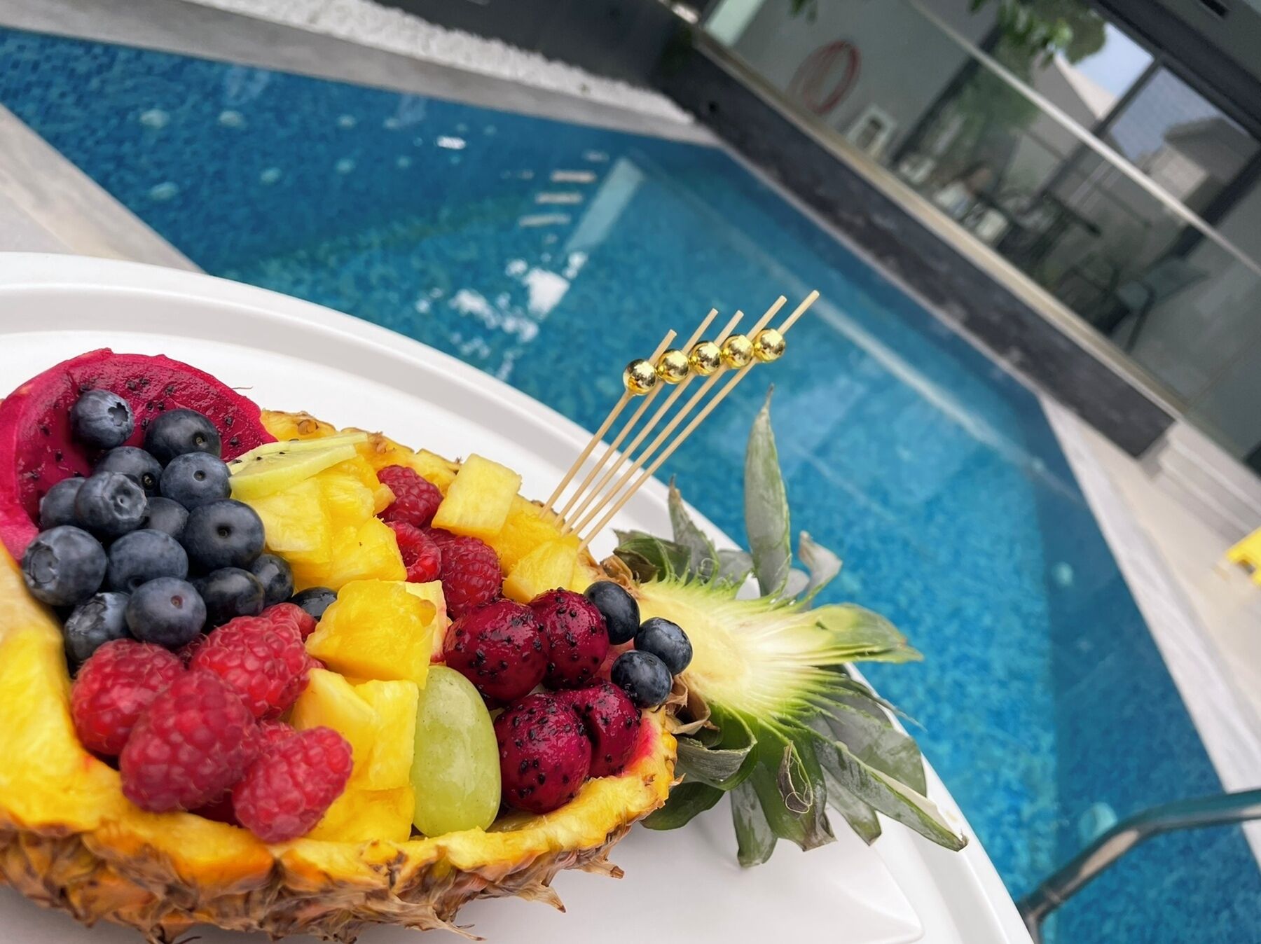 A fruit platter with a pineapple, blueberries, raspberries, and grapes, placed on a white plate by a poolside.