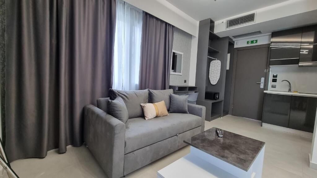 Una sala de estar moderna con un sofá gris, una mesa de centro y una cocina con electrodomésticos de acero inoxidable.