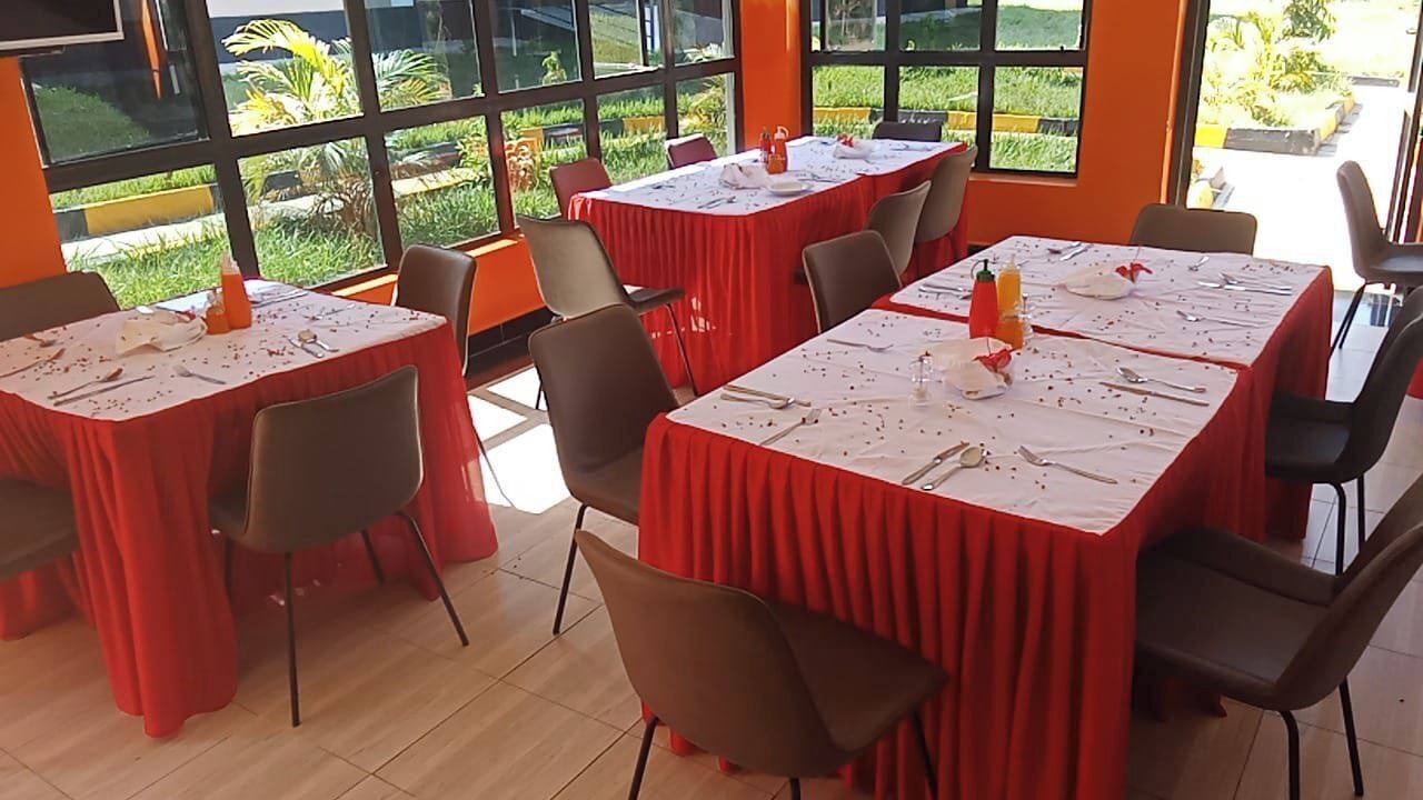A dining area with tables set for six, each covered with a red tablecloth. The room has large windows offering a view of greenery outside.