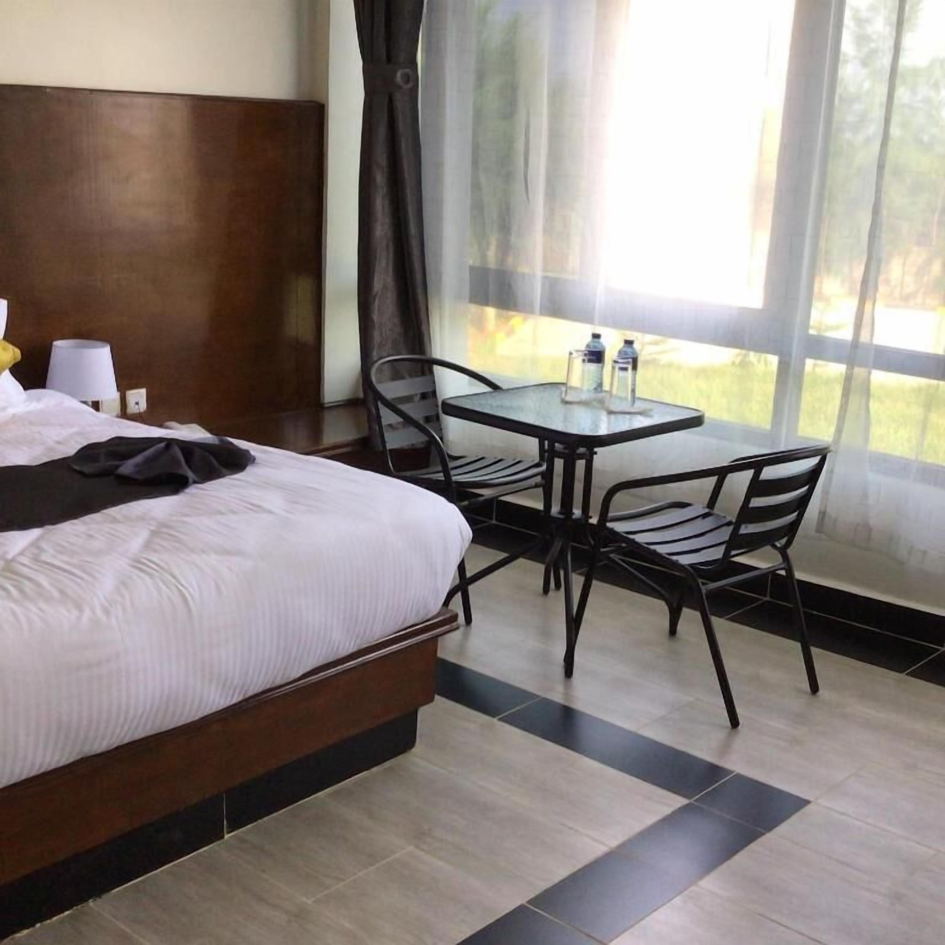 Habitación de hotel con cama, una pequeña mesa con sillas, ventana con cortinas y vistas a zonas verdes.