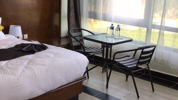 A hotel room with a bed, a small table with chairs, a window with curtains, and a view of greenery outside.