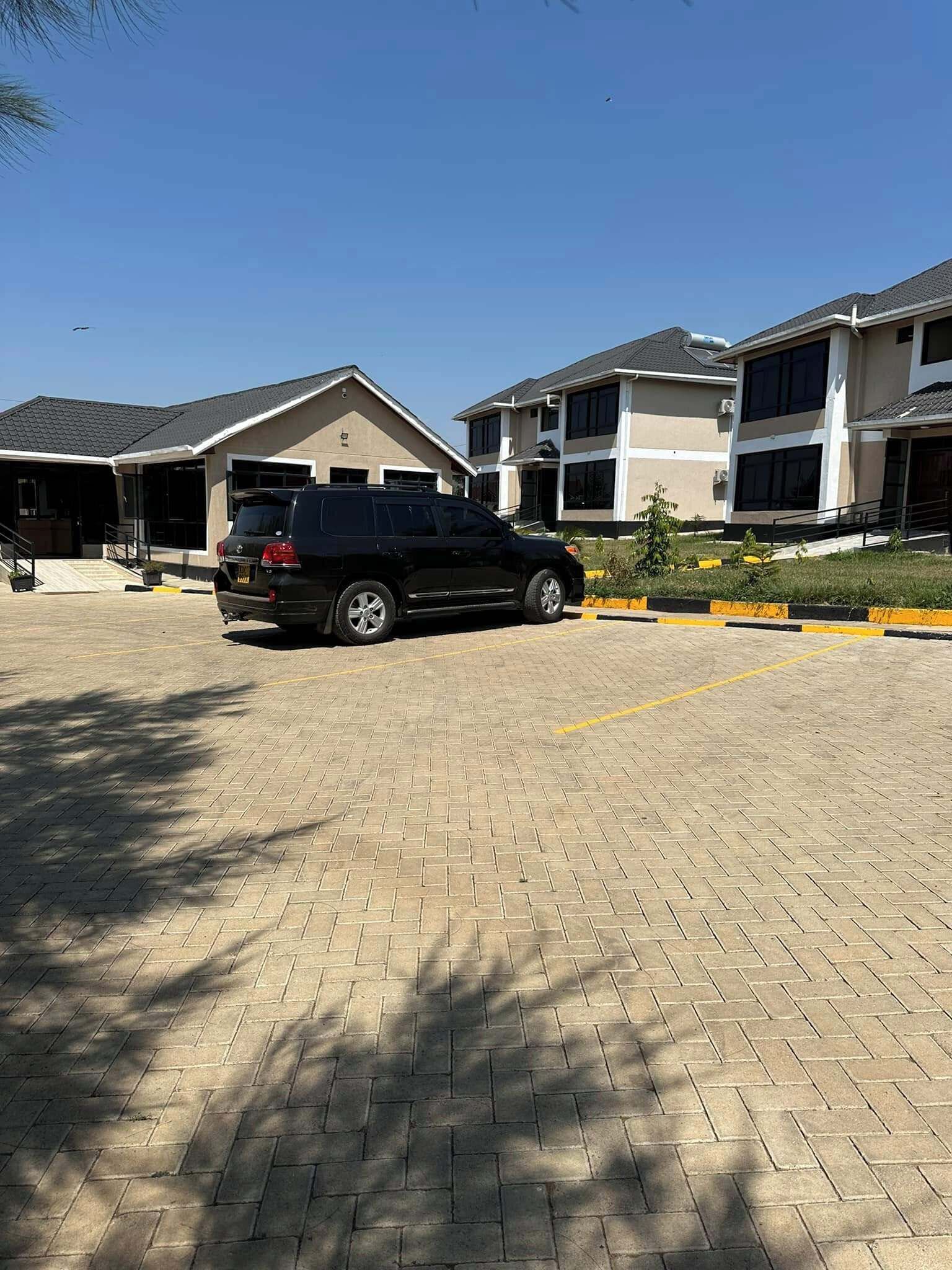 A parking area with a black SUV, paved with interlocking bricks, and surrounded by modern two-story buildings with large windows and a flat roof.