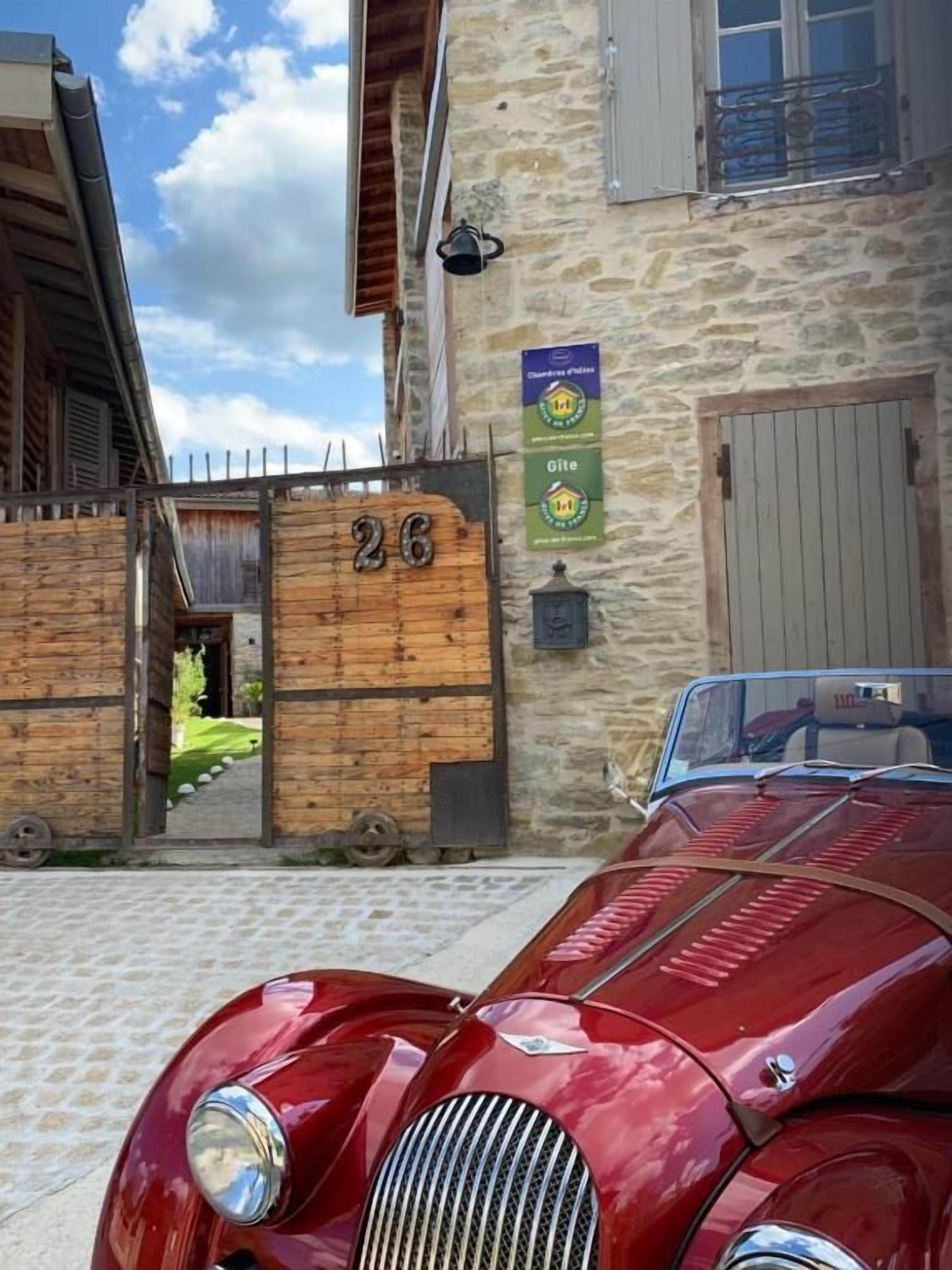 Un coche rojo antiguo estacionado frente a un edificio de piedra con un portón de madera que indica '26'.