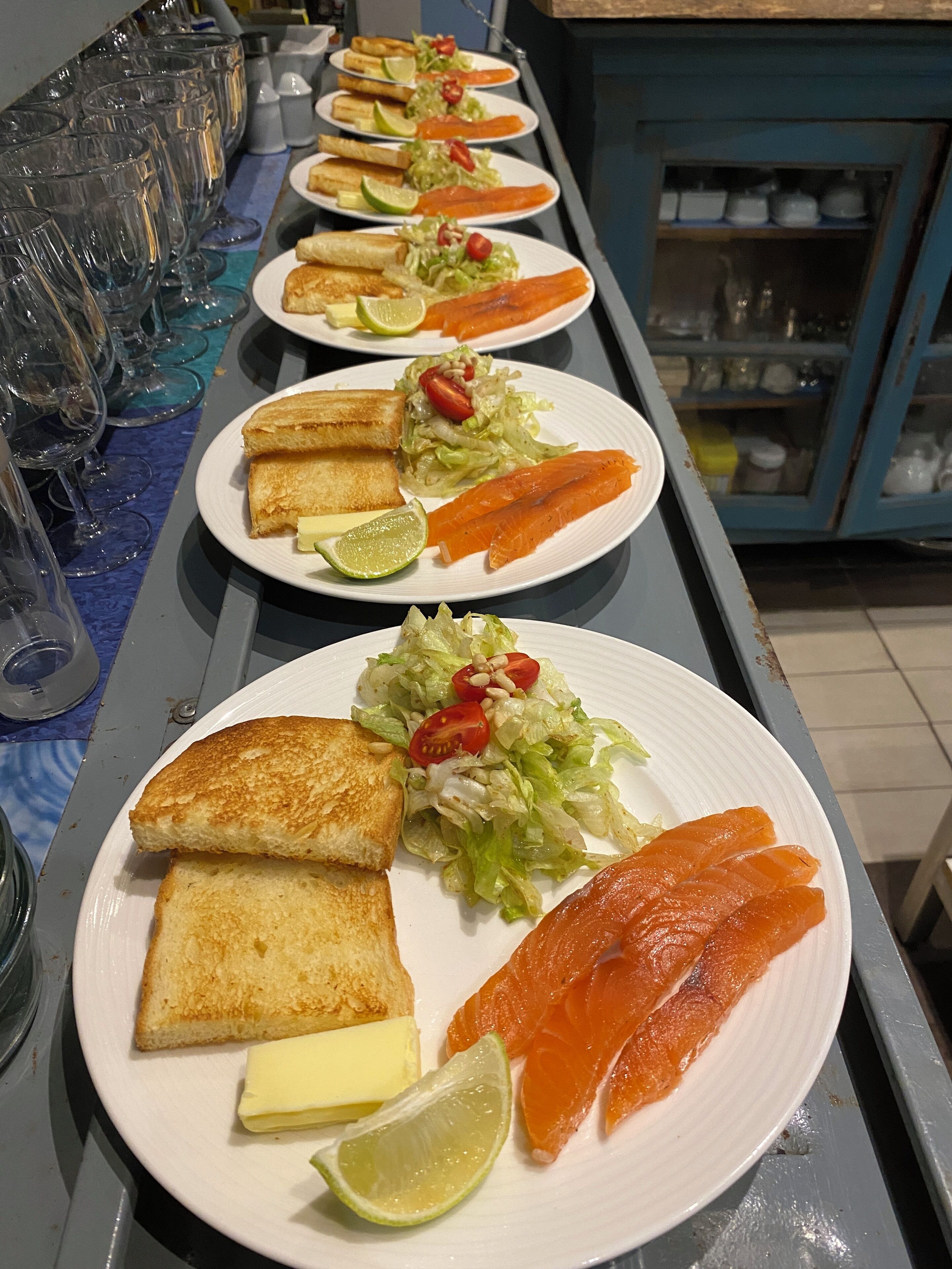 A buffet table with plates of food, including salmon, toast, and salad, and a row of empty glasses.