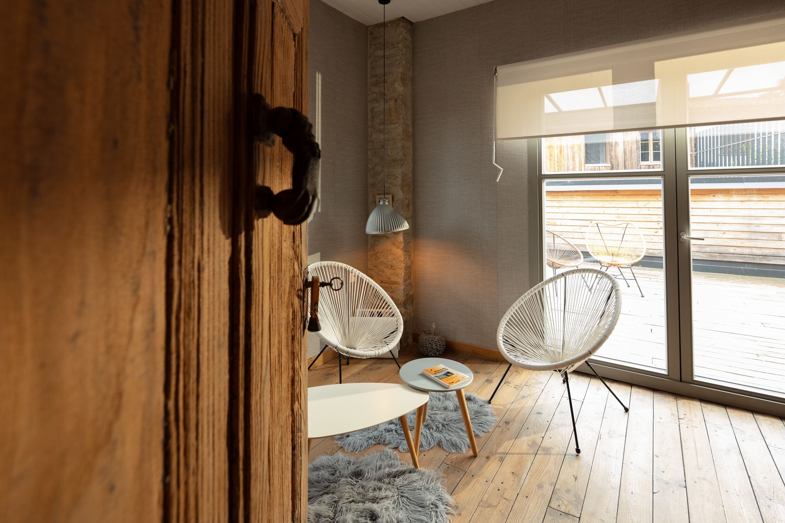 Un interior moderno con una puerta de madera, sillas de mimbre blanco, una pequeña mesa redonda y un ventanal con vistas al exterior.