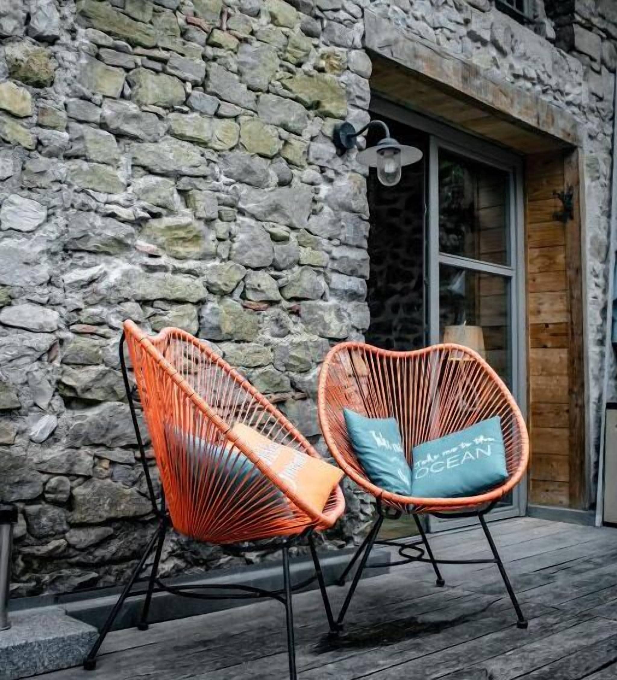 Deux fauteuils en osier avec des coussins sont placés à l’extérieur, devant un bâtiment en pierre doté d’une porte en bois et d’une fenêtre.