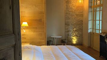 Un dormitorio con una cama grande, vigas de madera en el techo, paredes de piedra, una mesita y una ventana.