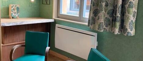 Ein Zimmer mit Türkis-Stühlen, einem Holztisch, einem Heizkörper und einem Fenster mit einem gardinierten Vorhang mit Blattmuster.