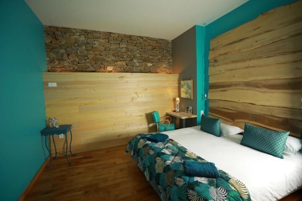 Une chambre avec un mur en pierre, un lambris en bois, un lit recouvert d’une couvre-lit à motifs de feuilles, une petite table et une chaise.