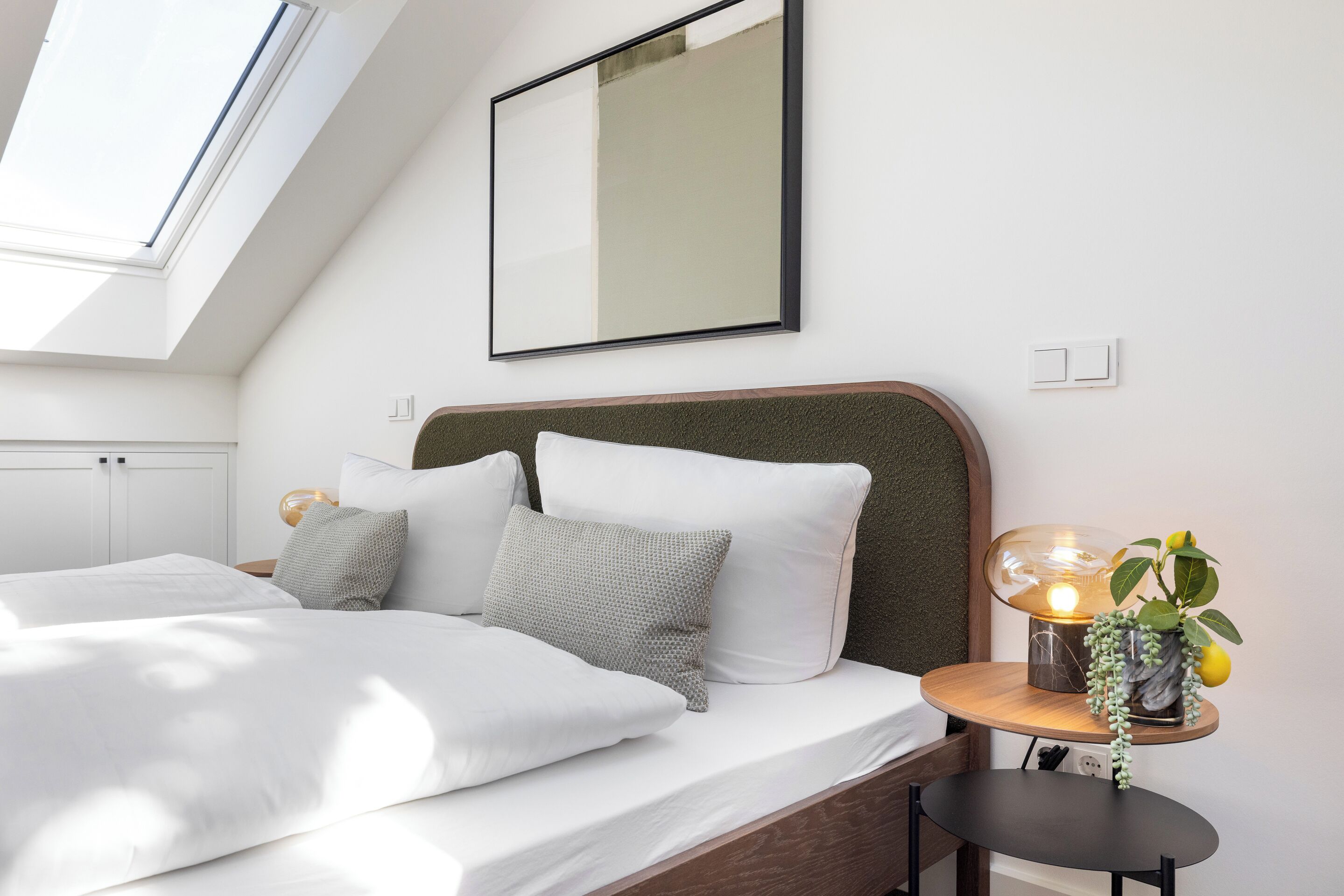 Une chambre avec un puits de lumière, un lit avec une literie blanche, une table de chevet ronde avec une lampe et un miroir fixé au mur.