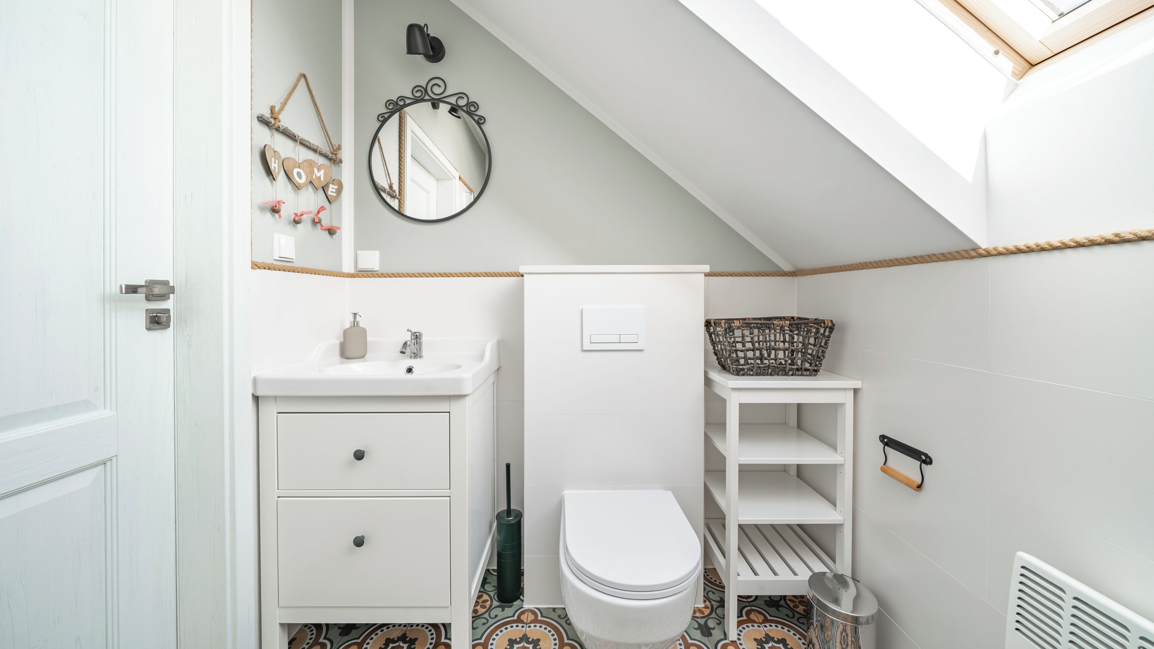 Et lille badeværelse med et hvidt håndvask, toilet og hylde. Et spejl hænger på væggen, og en kurv står på hylden.