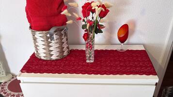 Een witte dressoir met een rode doek erop, een gevlochten mand, een vaas met bloemen en een rood wijnglas.