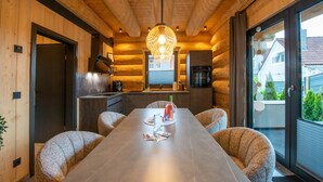 A modern log cabin interior with a dining area featuring a long table, comfortable seating, and a large window offering a view of the outside.