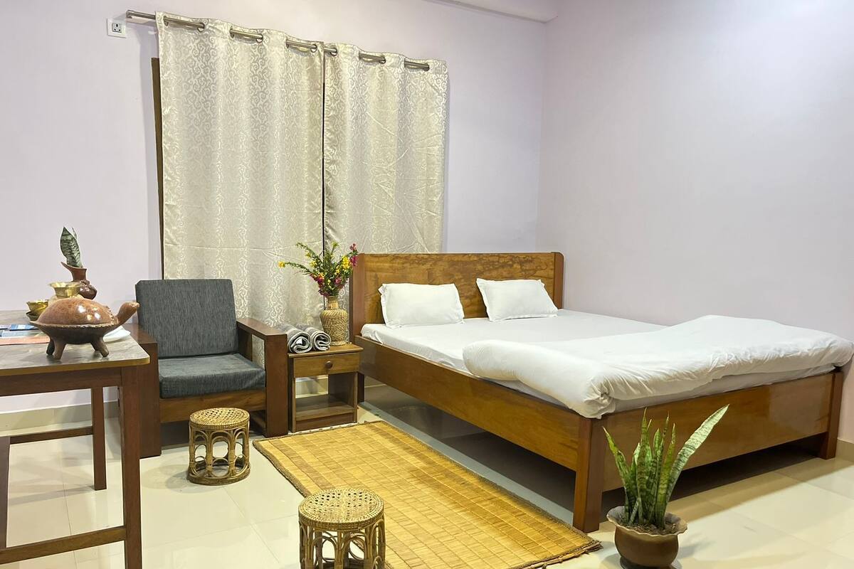 A hotel room with a wooden bed, a grey chair, a small table with a decorative turtle, a woven mat, and a potted plant.