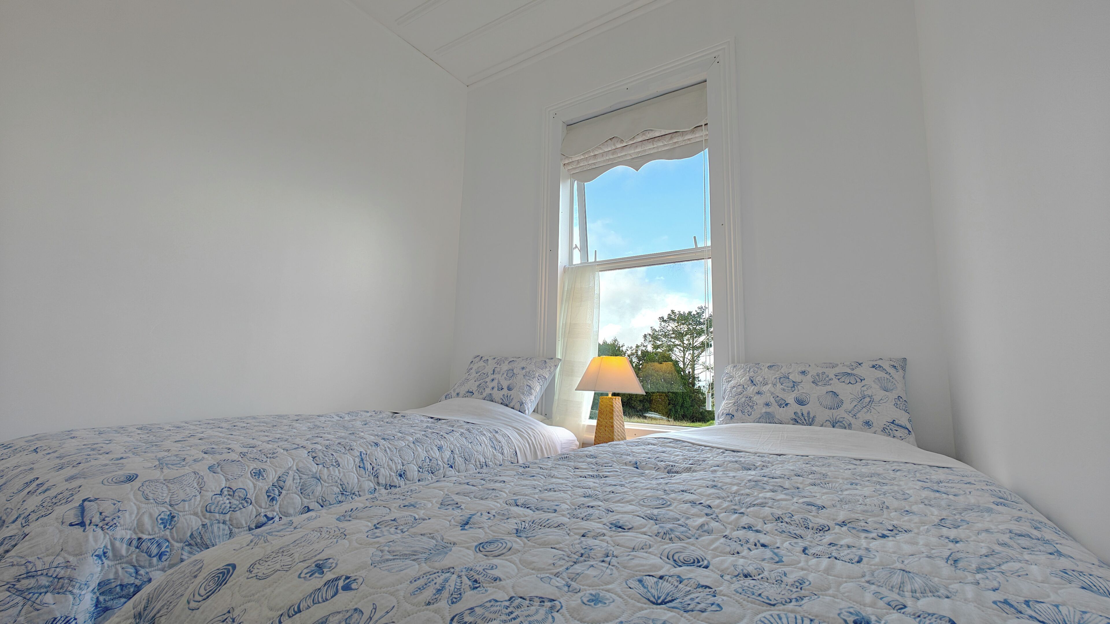 A bedroom with two beds, a window with a view of greenery, and a bedside lamp.