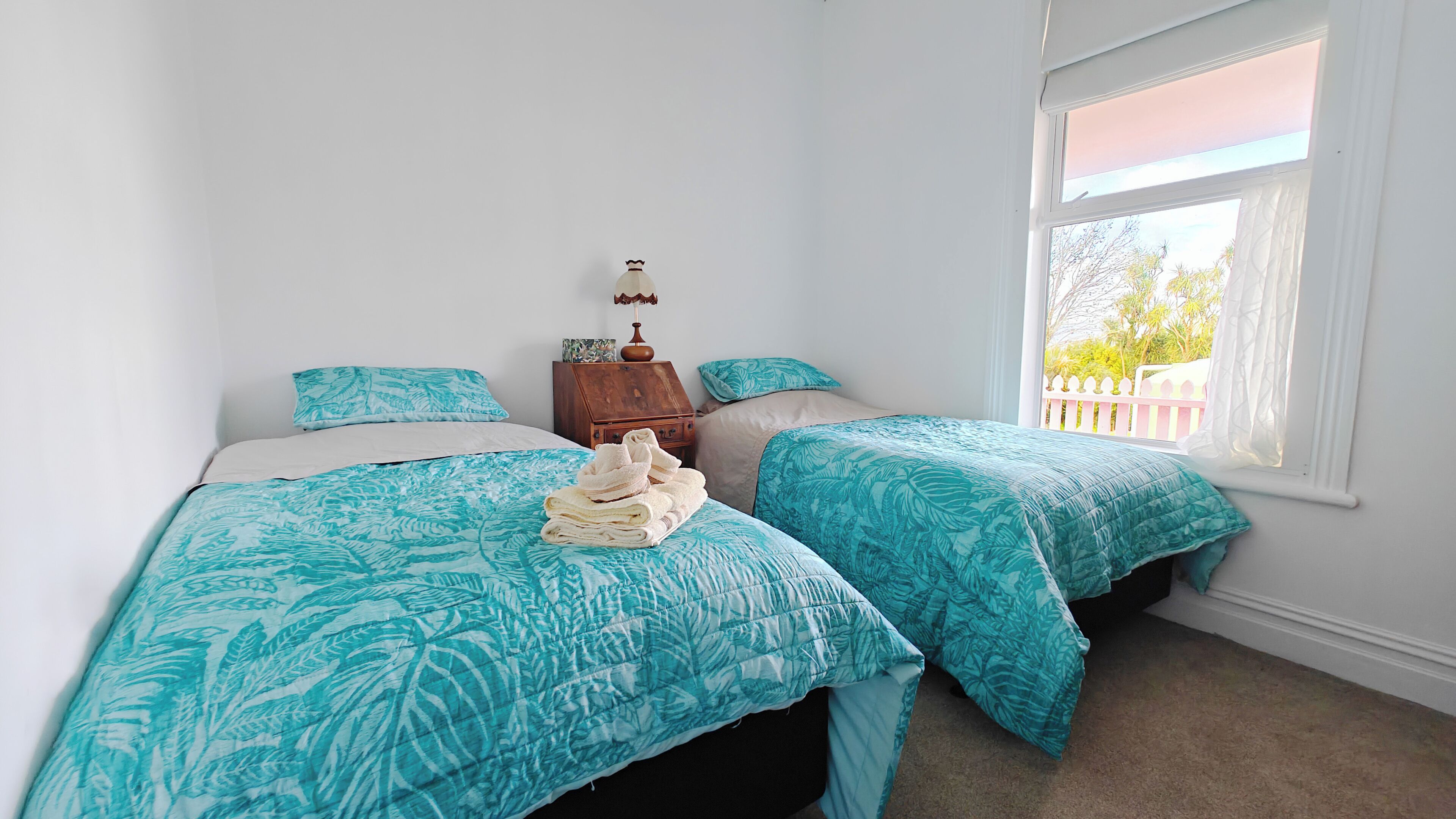 Quarto com duas camas, cada uma com colcha azul estampada com folhas, uma mesinha de cabeceira de madeira com abajur, e janela com vista para o verde lá fora.
