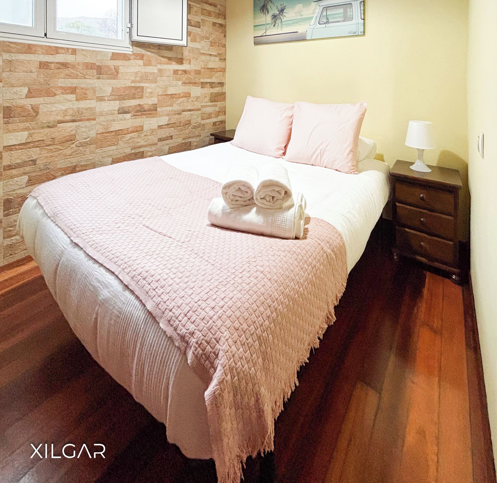 A bedroom with a stone wall, a bed with a pink blanket, two rolled towels, a nightstand with a lamp, and a painting of a vehicle on the wall.