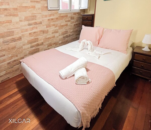 Una cama bien hecha con una manta rosa y toallas blancas, una mesita de noche de madera con una lámpara, y una pared con diseño de ladrillo.