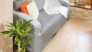 Un sofá gris con cojines naranja y blanco, una planta en maceta verde y un caja de madera con la etiqueta "WOOD BOX" en el alféizar de una ventana.
