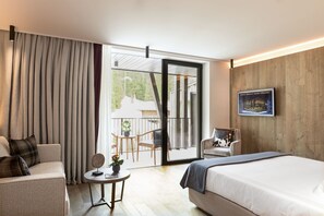 Une chambre d’hôtel avec un lit, un canapé, une petite table avec une plante et une chaise. Il y a un balcon avec vue sur l’extérieur.