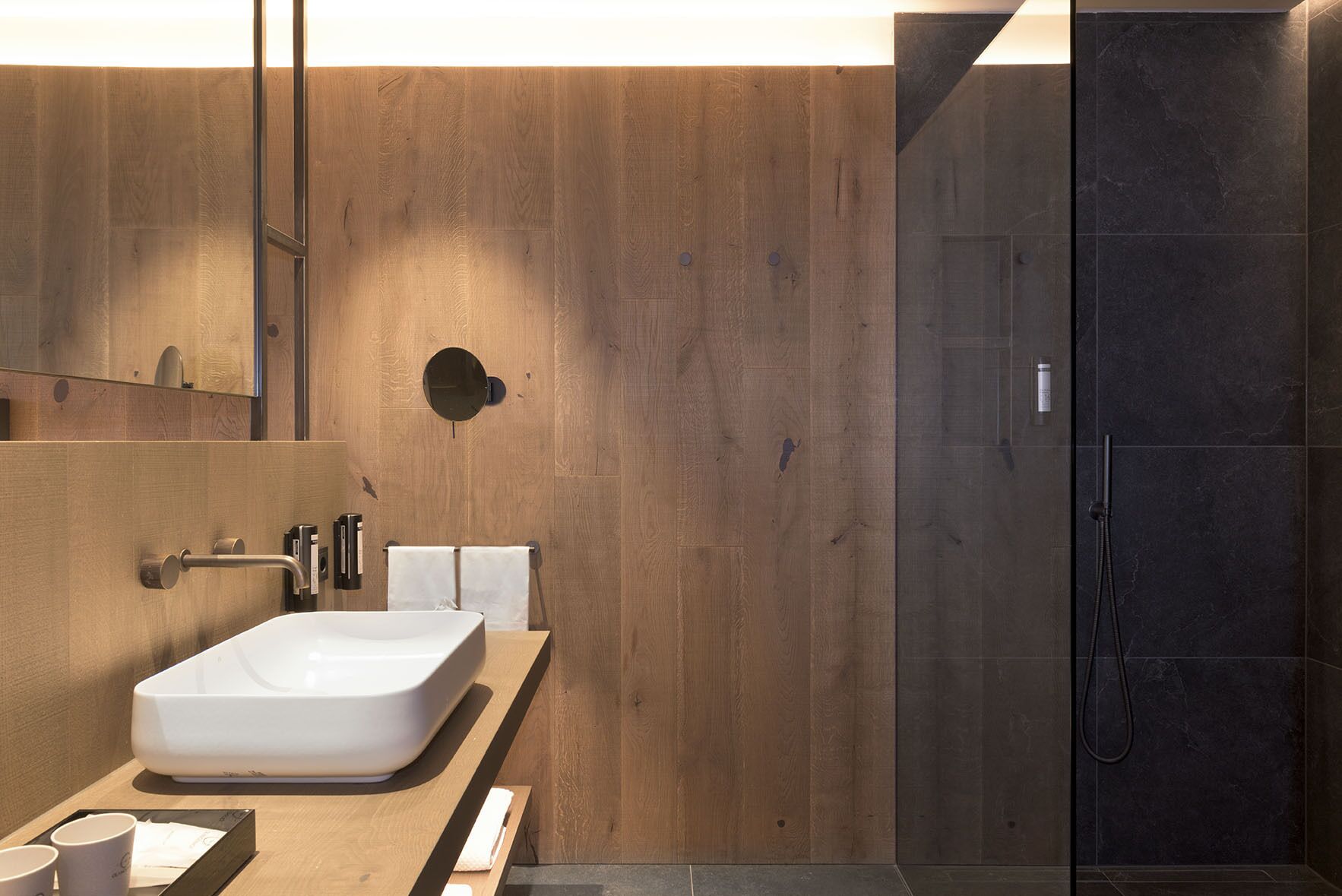 A modern bathroom with a wooden vanity, a large white sink, a mirror, and a dark shower area.