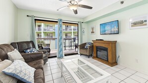 A living room with a ceiling fan, a fireplace, a television, and a view of a balcony with outdoor furniture. - Harbour Place 206 (Orange Beach)