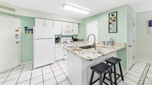 A kitchen with white cabinets, a white refrigerator, a microwave, and a sink. There are two black stools at the island. - Harbour Place 206 (Orange Beach)