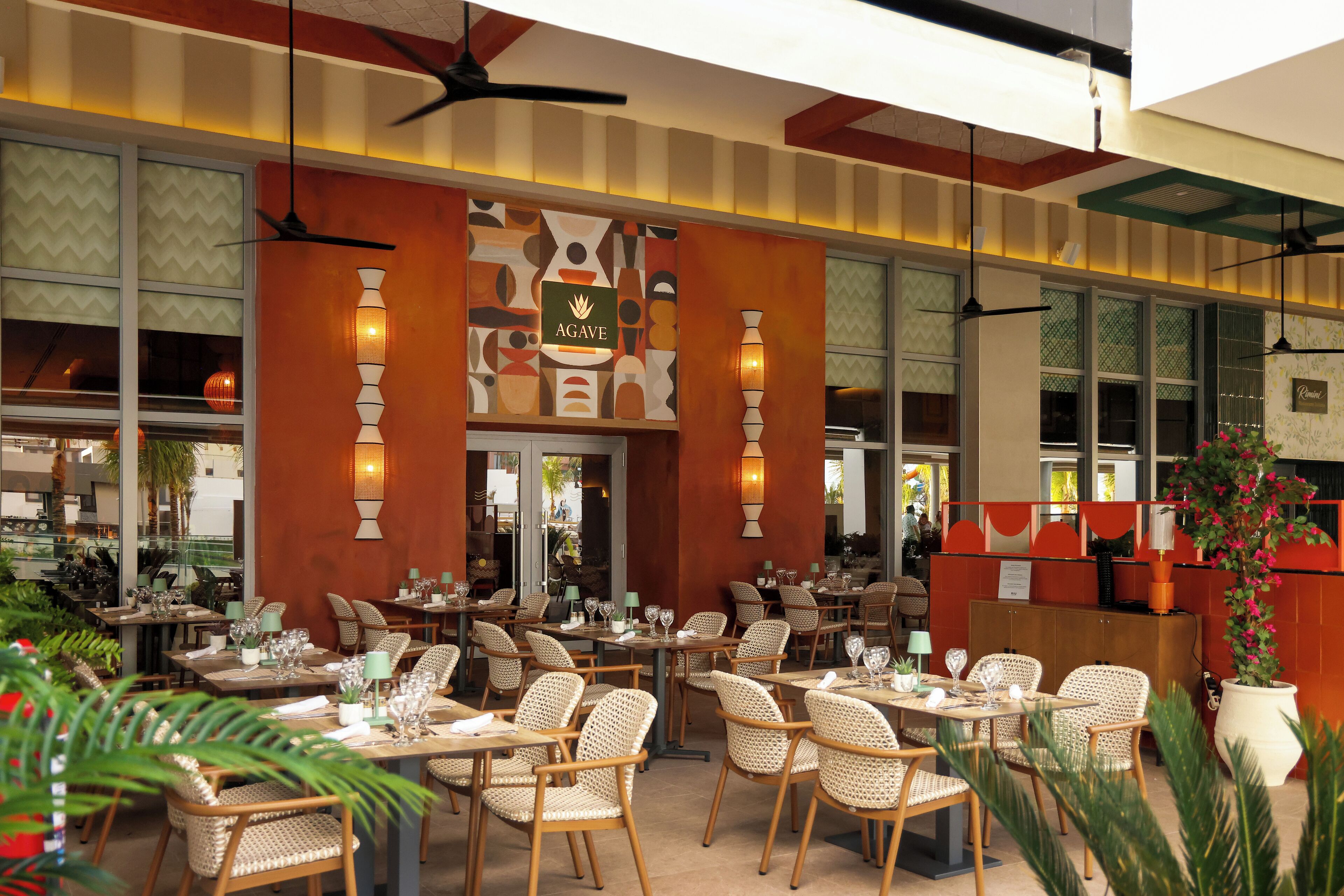 Un restaurante con sillas y mesas de mimbre, un letrero que dice "AGAVE" y ventanas grandes con diseños decorativos.
