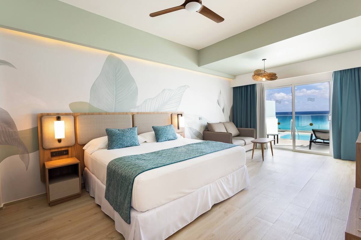 Una habitación de hotel moderna con una cama grande, una mesita de noche, una zona de estar con vista al océano y un ventilador de techo.