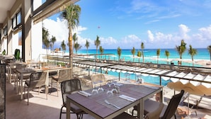Un área de comedor en la azotea con mesas y sillas, con vistas a una piscina y una playa con palmeras.