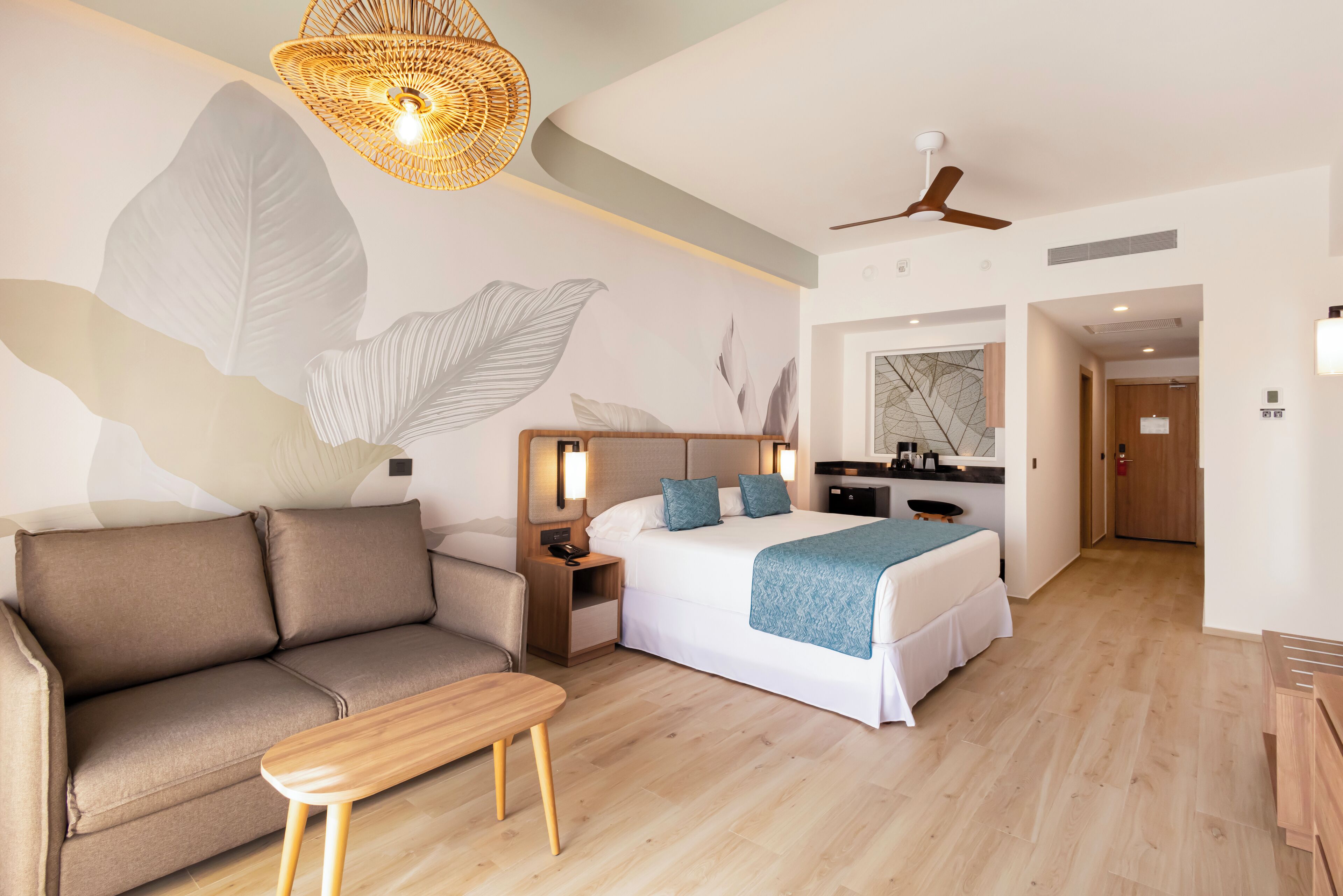 A modern hotel room with a large bed, a sofa, a small table, a desk, and a kitchenette.