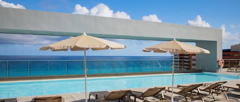 Zona de piscina con sillones de descanso y sombrillas, con vista a un océano azul y despejado.